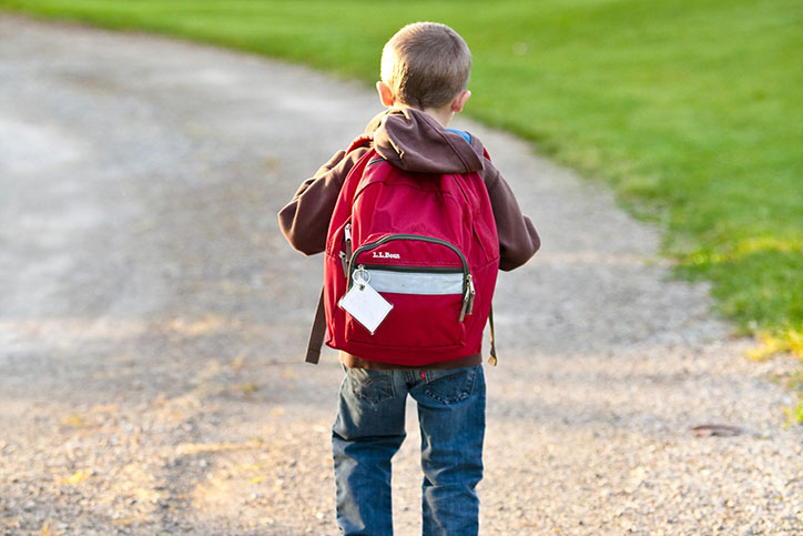 Young child walks down gravel path with red backpack.