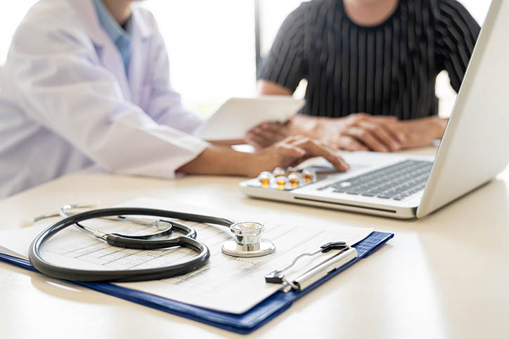 Bottom half of a photo showing a doctor and patient in a consultation. There is a clipboard, stethoscope, medicine, and a laptop on the desk.