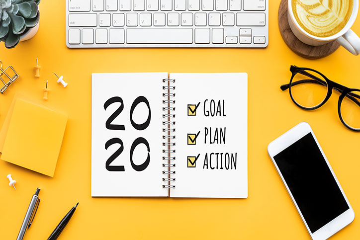 Yellow desk with notepad written 2020 and goals plans and actions. Other desk items surround the notepad including pens, thumbtacks, sticky notes, a plant, a keyboard, a cup of coffee, glasses, and a cellphone.