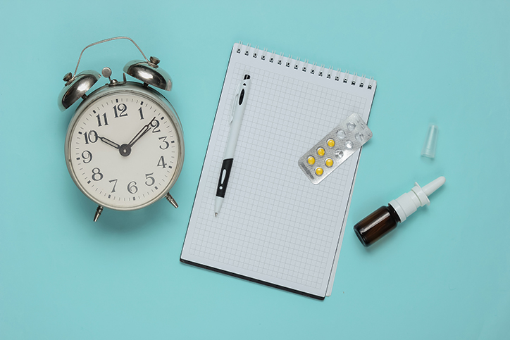 Medical still life on a blue studio background. Top view. Flat lay. Items include a clock, notepad, pill pack, and nasal spray.
