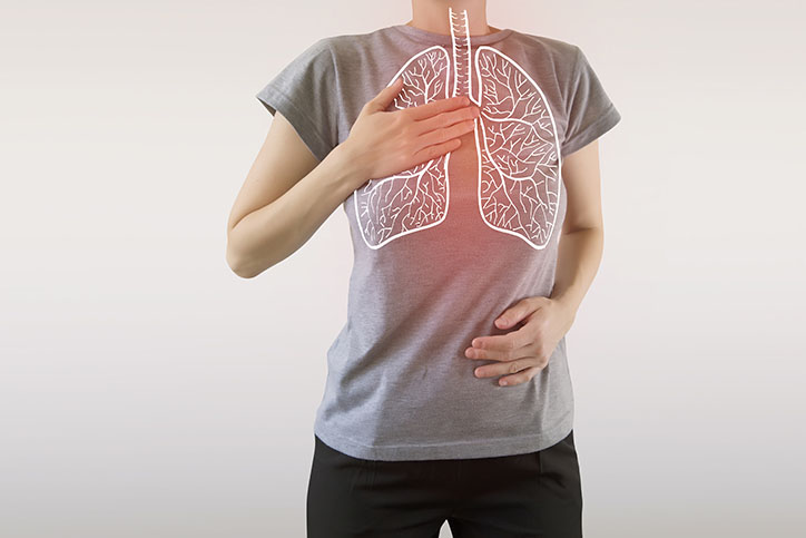 person in a gray shirt holding their chest and stomach with their hands. There is a white outline of the lungs with a red glow behind it.