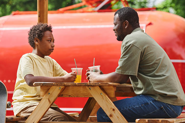 Dad talking to his son sitting at table during their rest at outdoor cafe with cold juice