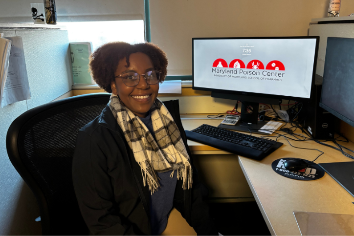 A photo of Dr. Kayla Bourgeois sitting at a desk with computer in the background with a screen saver with the Maryland Poison Center logo on it