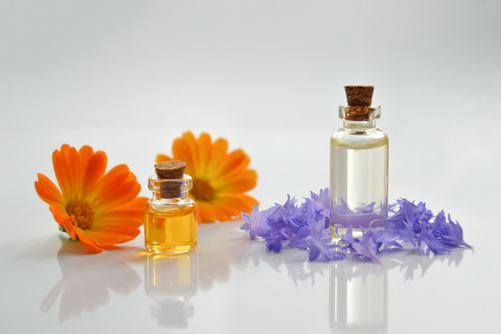 Glass bottles of liquid with cork stoppers displayed beside orange and purple flowers on a white background.