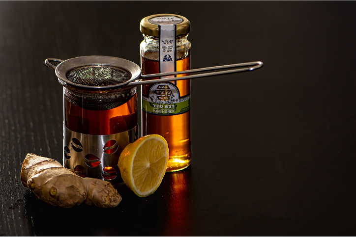A jar of golden honey, a metal tea infuser resting across a glass of dark amber tea, a half lemon, and a piece of fresh ginger root arranged on a dark tabletop.